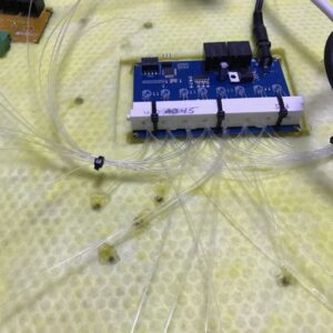 IntegratedLEDEngine - WDM Lighting Blue circuit board mounted on a white holder with multiple connections, surrounded by a chaotic bundle of clear fiber optic cables on a yellow lab surface.